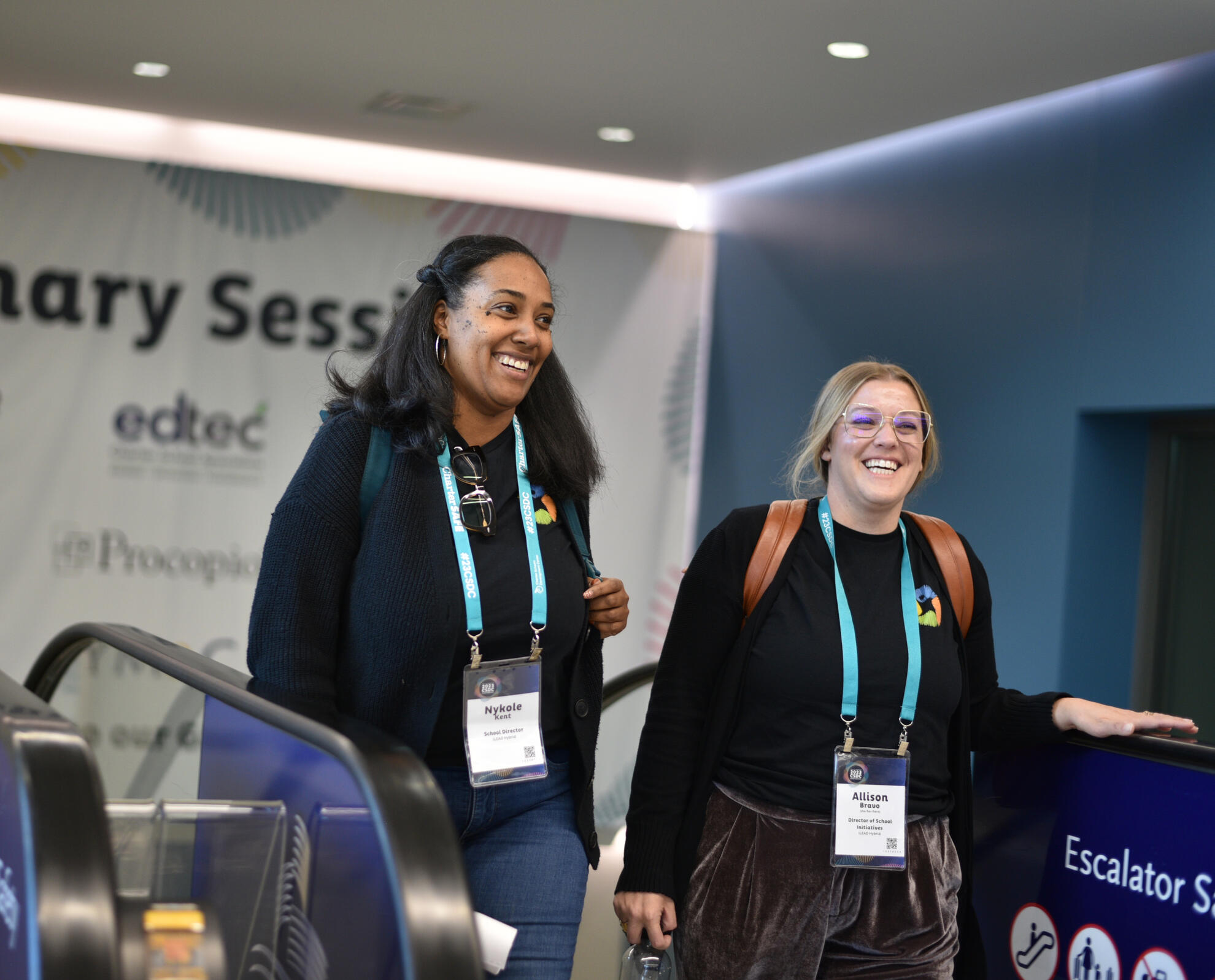 Two women exiting a plenary session with lanyards and conference badges, candidly photographed mid-escalator ride. Captured with warmth and movement, the image reflects ease, professionalism, and post-session energy.