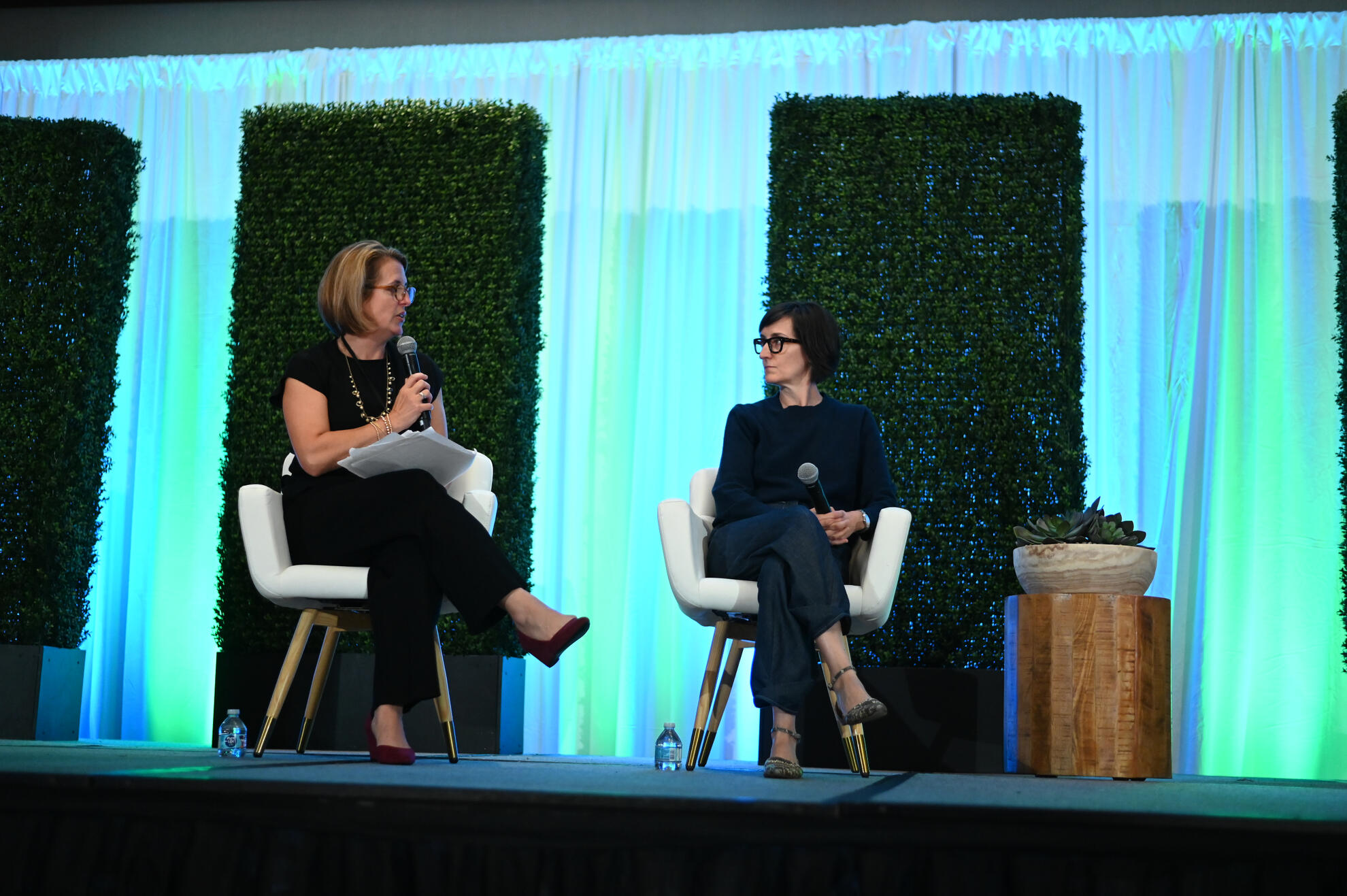 Two women seated on stage in a panel discussion, framed by greenery and soft drapery. A clean, polished conference setup captured with a documentary eye, highlighting leadership and thoughtful exchange.
