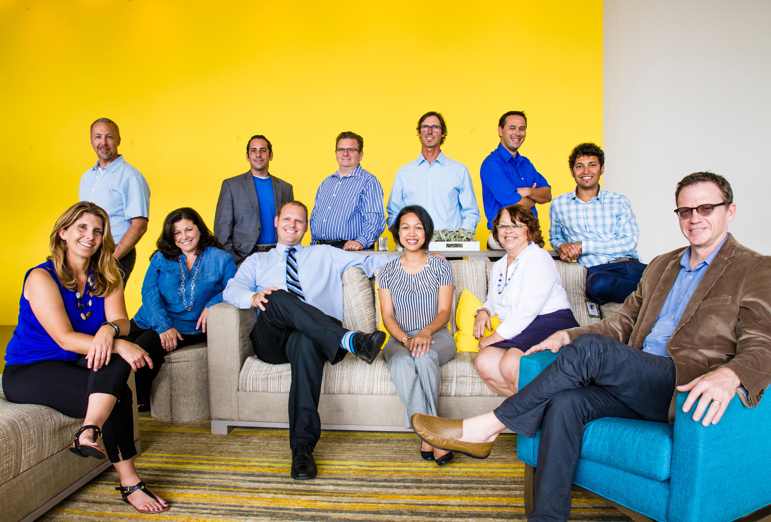 Team portrait of twelve professionals seated and standing against a bright yellow backdrop, blending formal and casual energy in a dynamic, color-coordinated group composition.